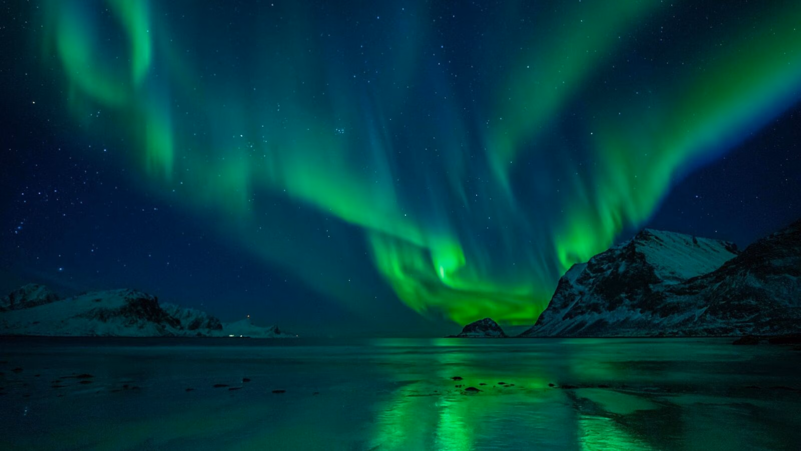 Green northern lights illuminate a starry night sky above snowy mountains and a reflective body of water.