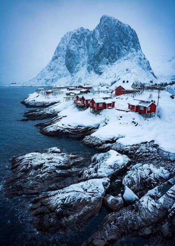 Red wooden houses on a snowy coastline in front of a massive, steep mountain peak under a cloudy sky.