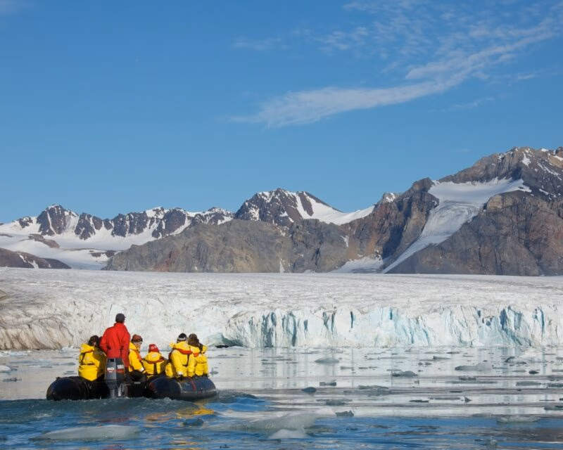 People in an inflatable boat navigate icy waters in front of a large glacier and jagged mountains.