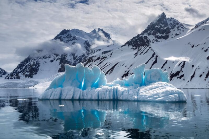 A large, translucent blue iceberg floating in calm water with snow-covered mountains in the background.