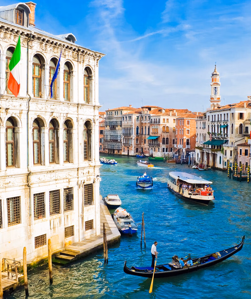 A gondolier navigates a black gondola on a busy blue canal in Venice, lined with historic Venetian buildings.