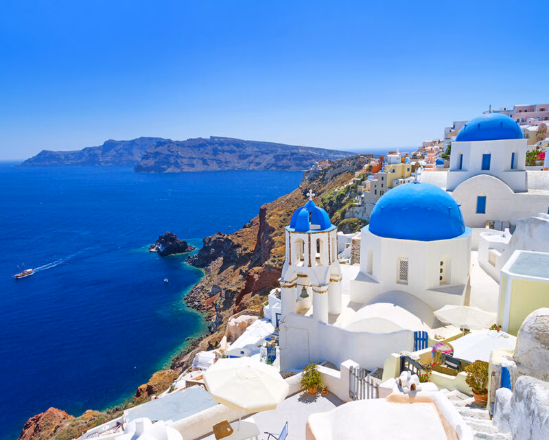 Whitewashed churches with bright blue domes overlook a rugged coastline and deep blue sea under a clear sky.