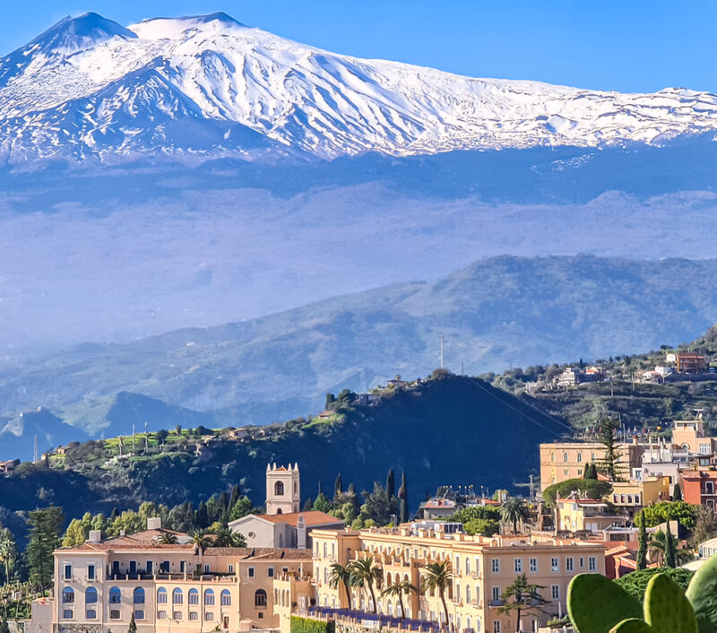 A hillside town with peach-colored buildings sits below a towering, snow-covered mountain under a bright blue sky.