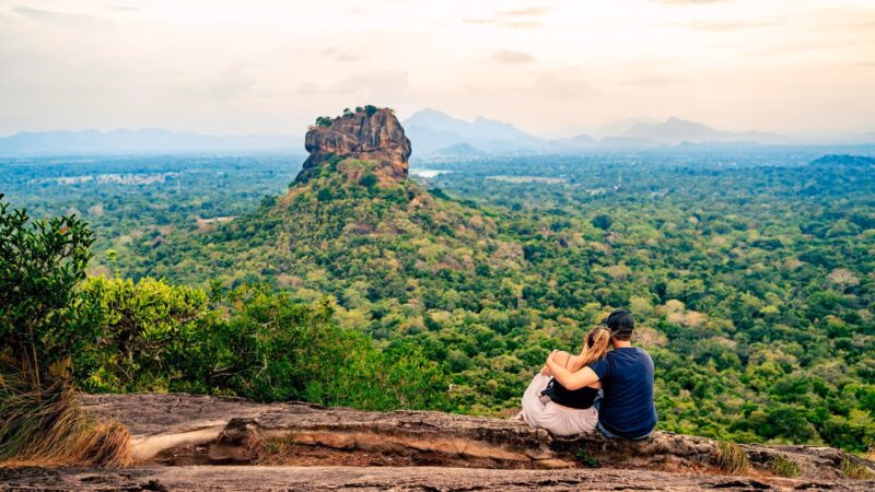 A couple sits on a ledge overlooking a vast forest toward Sigiriya rock.