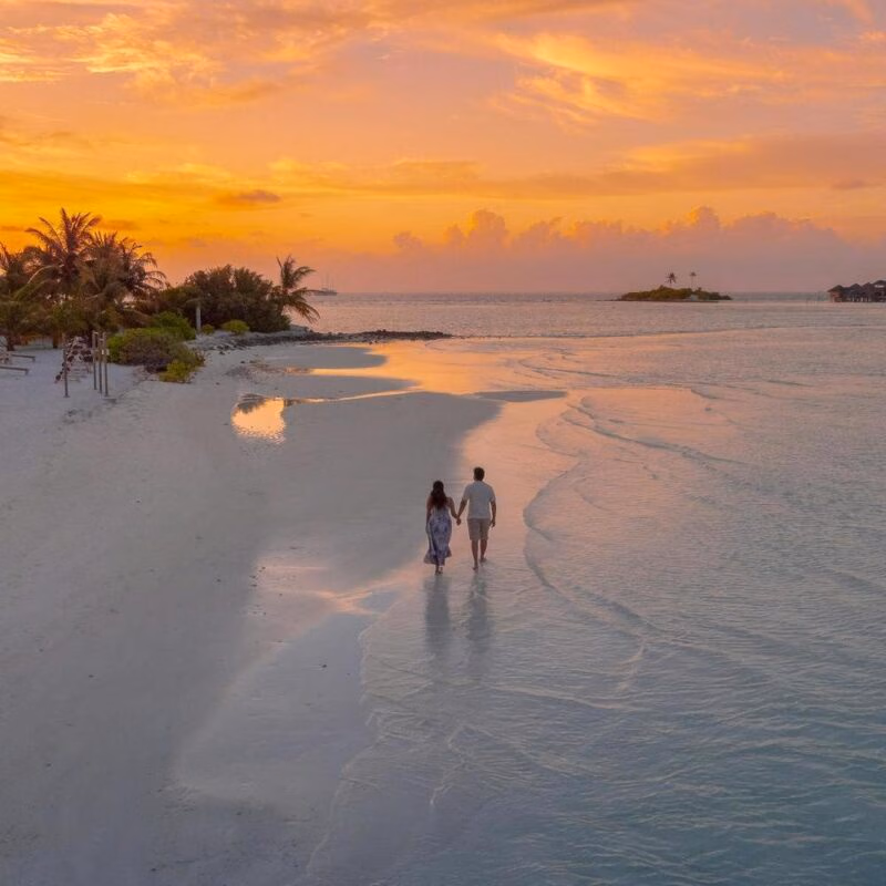 A couple walks along the edge of the ocean on a beach at sunset.