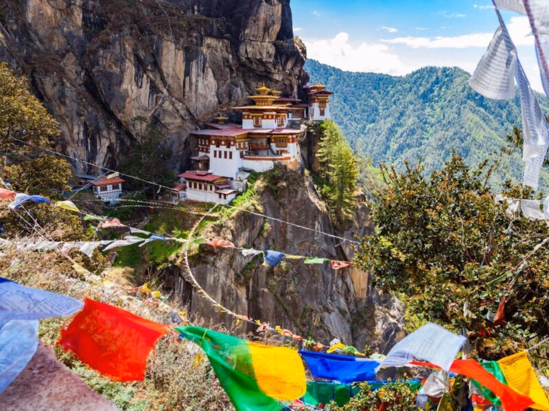 A cliffside monastery built into a steep rock face behind colorful hanging prayer flags.