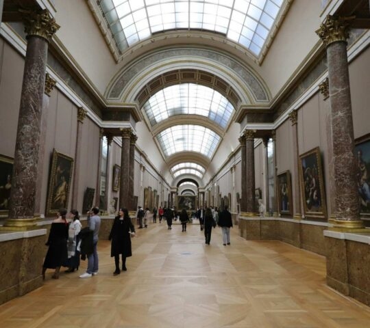 A hallway in the Louvre with glass ceiling and marble pillars, lined with large paintings with people admiring them