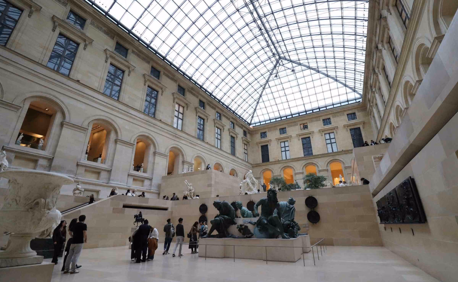 Tourists admiring a light stone gallery with glass roof filled with marble statues inside the Louvre, Paris