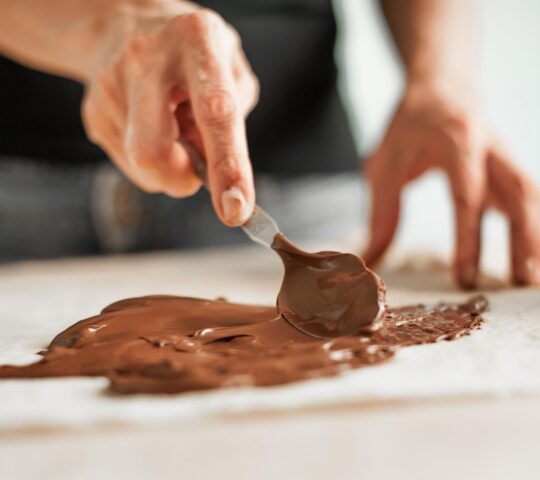 Close up of a man's hands using a spoon to smooth melted chocolate onto a board for chocolate making