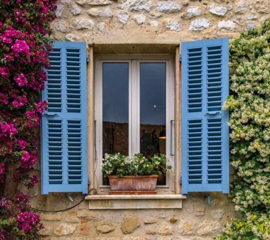 A blue shuttered window in a stone house surrounded by bougainvilia and jasmine flowers, Saint Paul de Vence, South of France