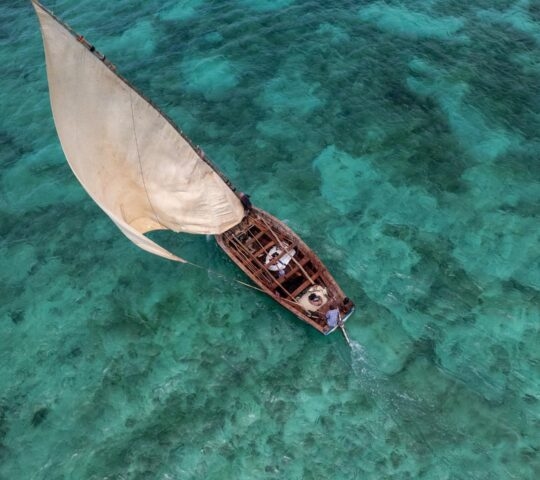 High-angle view of a wooden dhow boat with a white sail on vibrant turquoise water.