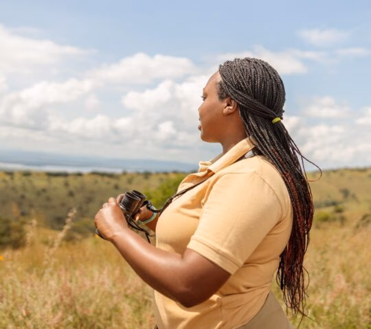 A woman holding binoculars looks out across an open, rolling green landscape under a blue sky with white clouds.