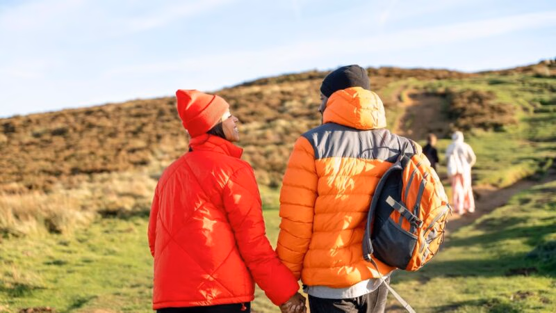 Rear view of a couple in red and orange jackets holding hands while hiking up a grassy hillside.