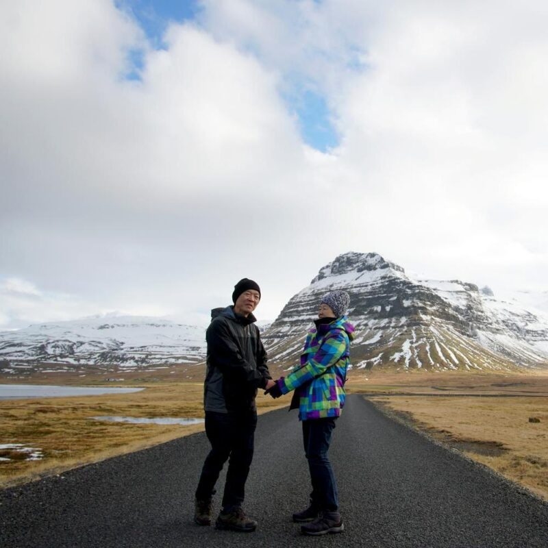 A couple holds hands on an empty road with a large snow-covered mountain in the background.