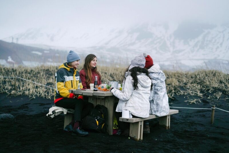 Four people in winter coats having a picnic at a wooden table on black sand with snowy mountains behind them.