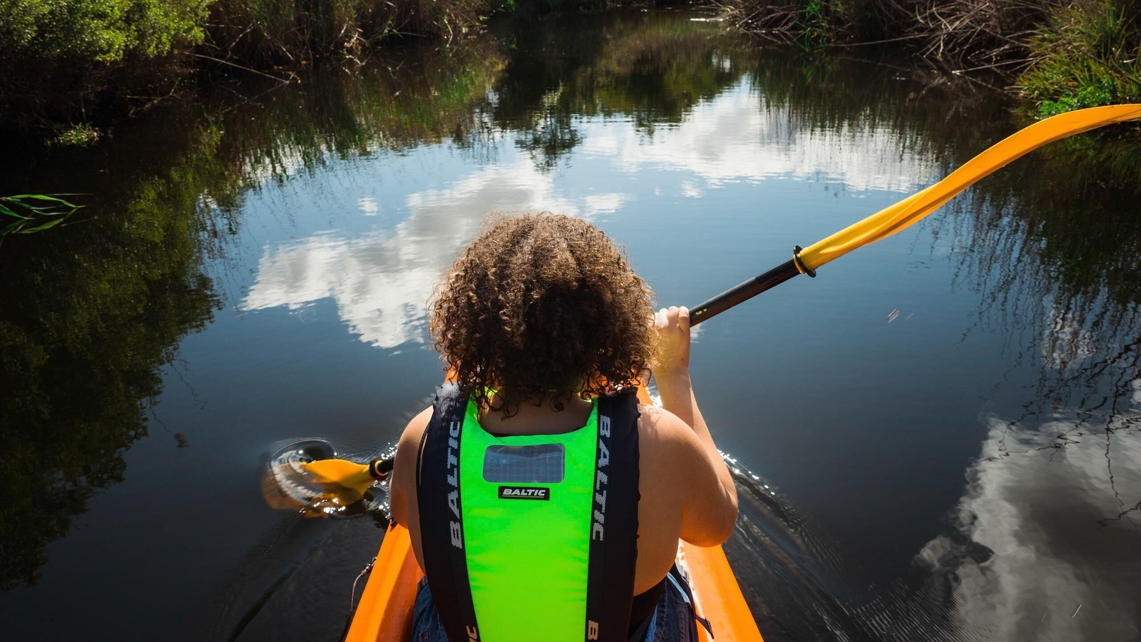 First-person rear view of a person with curly hair paddling a canoe down a still waterway under a bright sky.