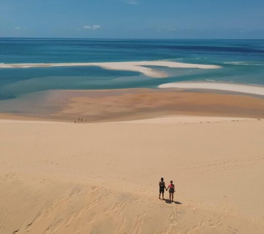 A couple holds hands on a large sand dune overlooking coastal lagoons and turquoise sea in the Bazaruto Archipelago.