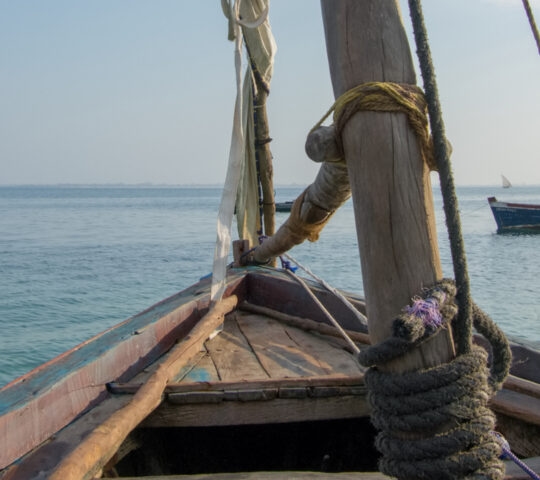 The wooden bow and rigging of a dhow boat sailing on clear, blue water under a hazy sky.