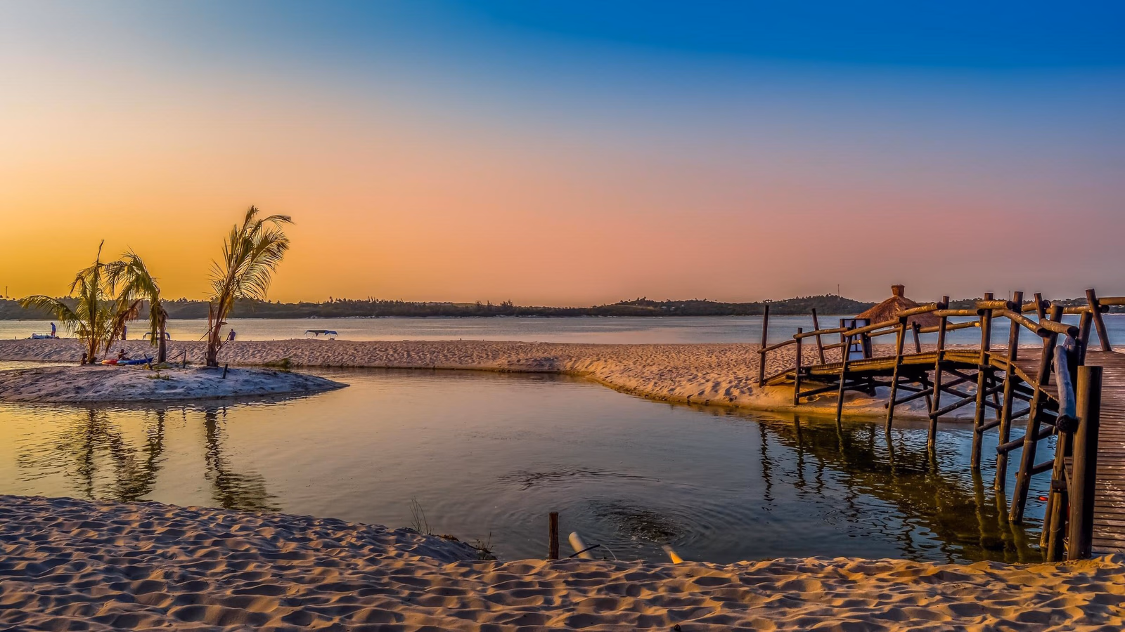 A wooden footbridge next to a beach lagoon with palm trees under a vibrant orange and blue gradient sunset sky.