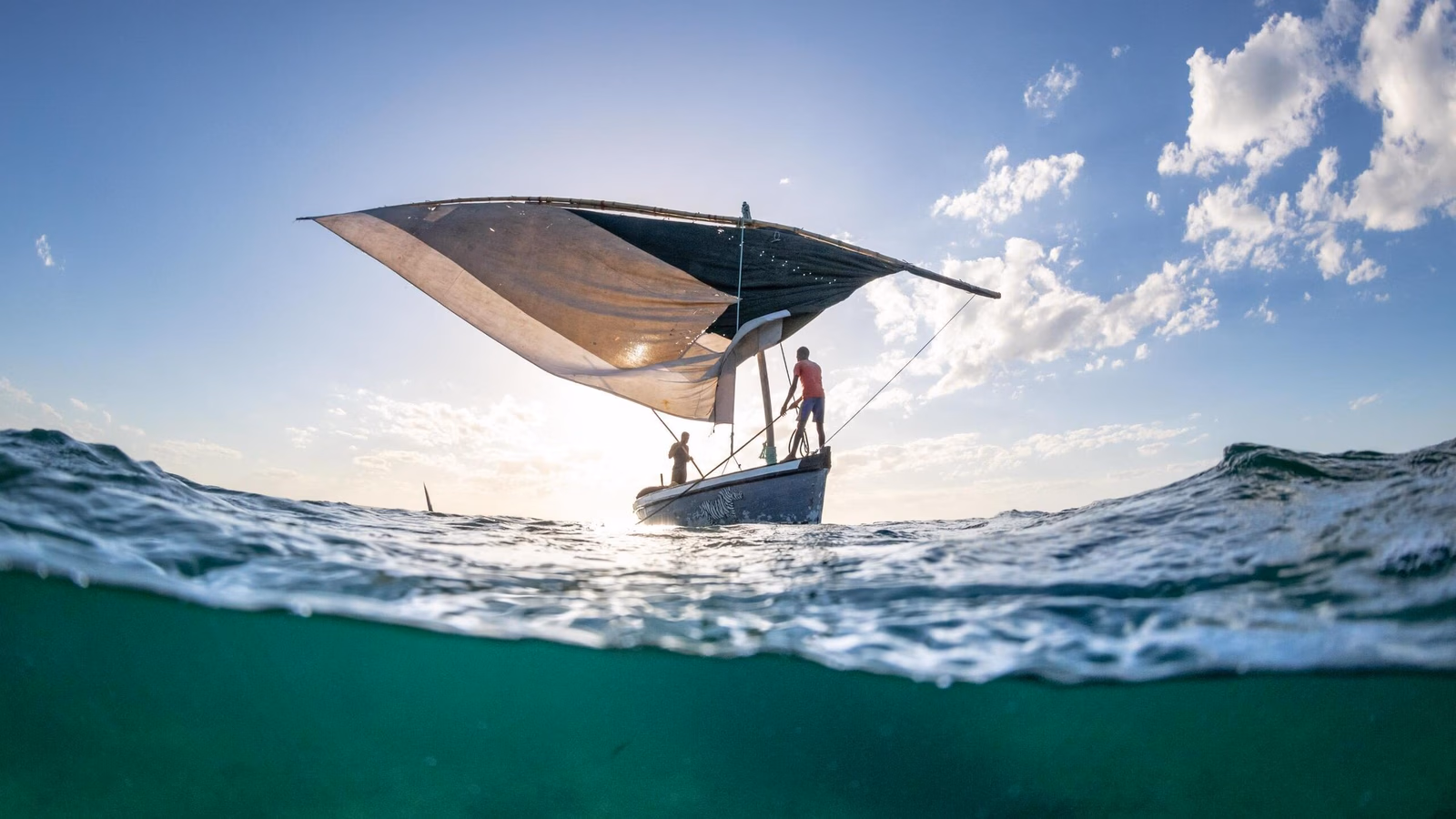 A split underwater and surface view of a traditional dhow boat sailing on clear turquoise ocean waters.