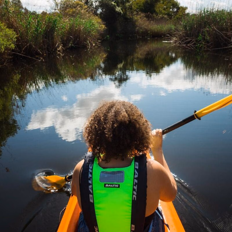 First-person rear view of a person with curly hair paddling a canoe down a still waterway under a bright sky.