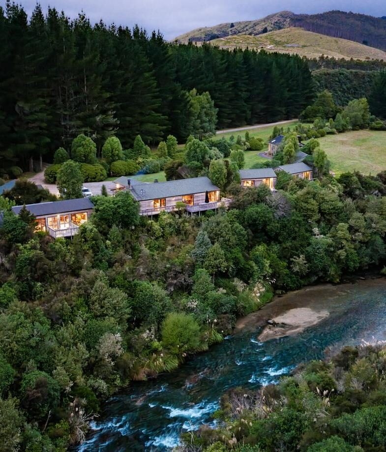 An aerial view of Poronui Lodge’s riverside cabins, followed by the peaceful pool terrace at Hapuku Lodge. Two secluded New Zealand stays surrounded by forest, water and mountain scenery.