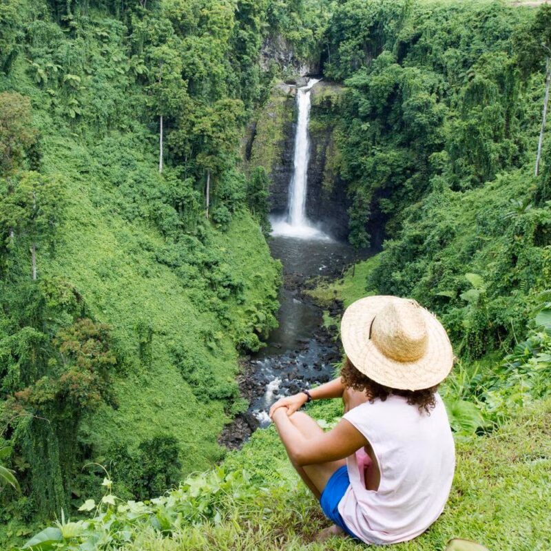 A person in a straw hat sits on a grassy cliff looking at a tall waterfall in a lush green valley.