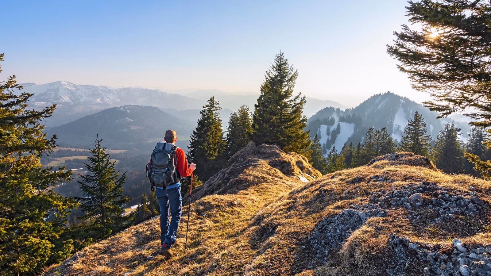 A hiker with a backpack walks on a mountain ridge overlooking a vast valley with evergreen trees and distant snowy peaks.