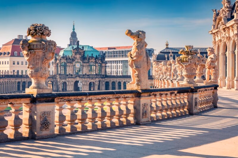 Classic stone statues and ornate urns line a grand terrace overlooking historic baroque buildings in Dresden, Germany.