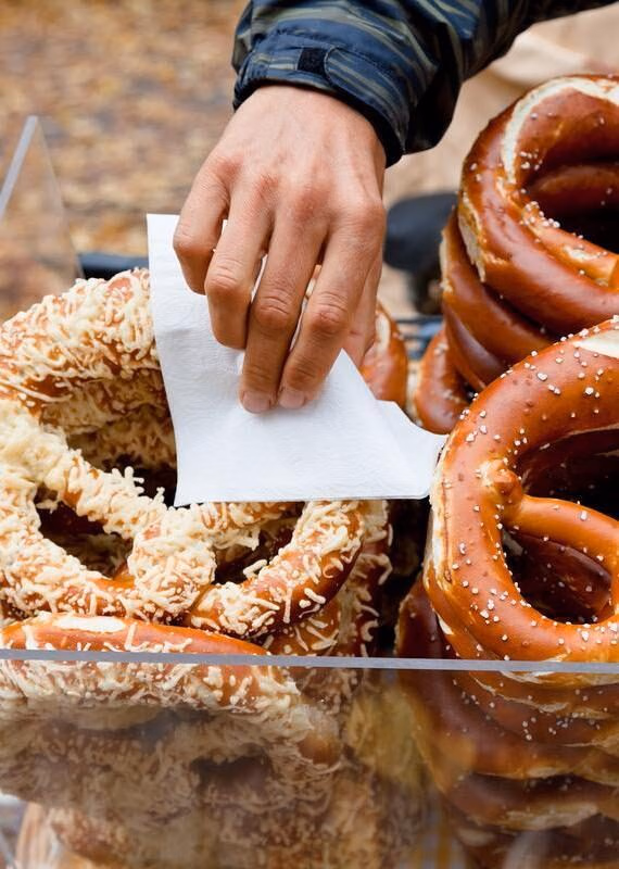 A hand holding a white napkin reaches for a cheese-topped pretzel next to stacks of salted pretzels behind glass.