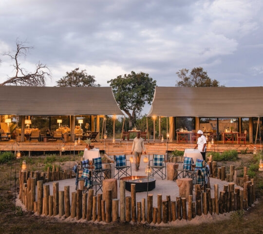 Exterior view of a tented lodge with open walls and a few trees in the background, and people around a fenced firepit in the foreground.