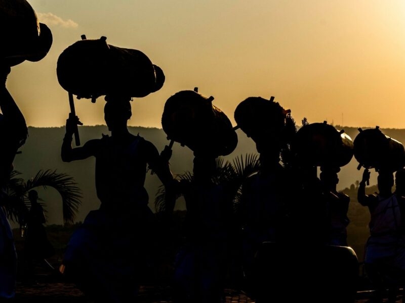 Silhouettes of dancers carrying large drums on their heads against a bright sunset sky.