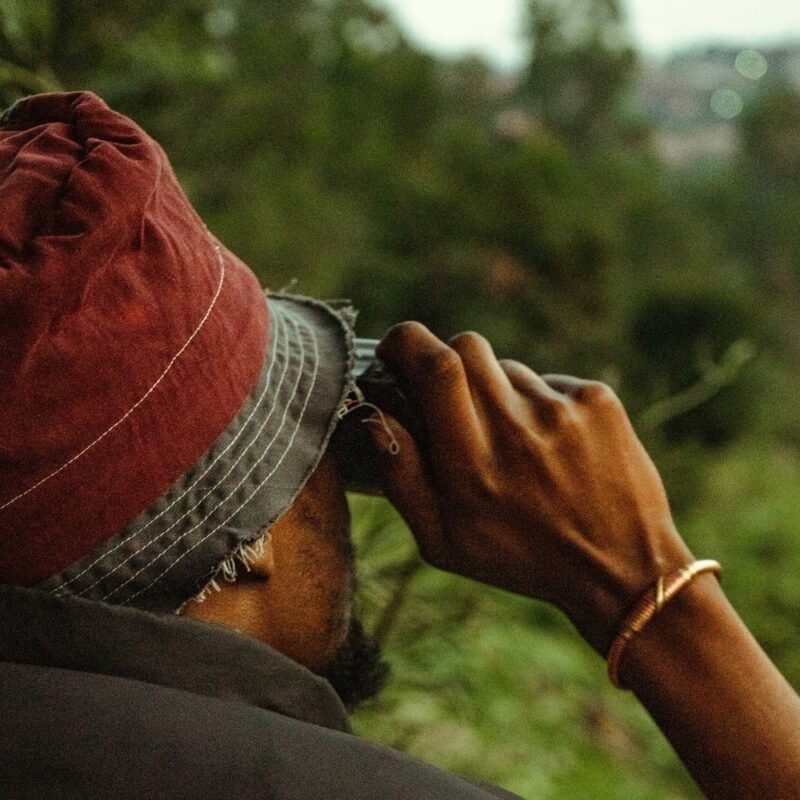 Close-up profile of a person in a bucket hat looking through binoculars at a green landscape.