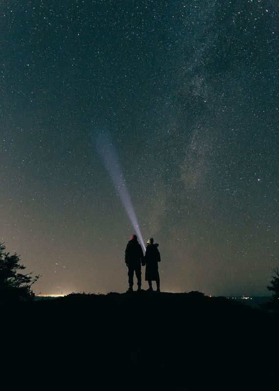 Silhouette of a couple standing on a hill under a vast, star-filled night sky with a flashlight beam.