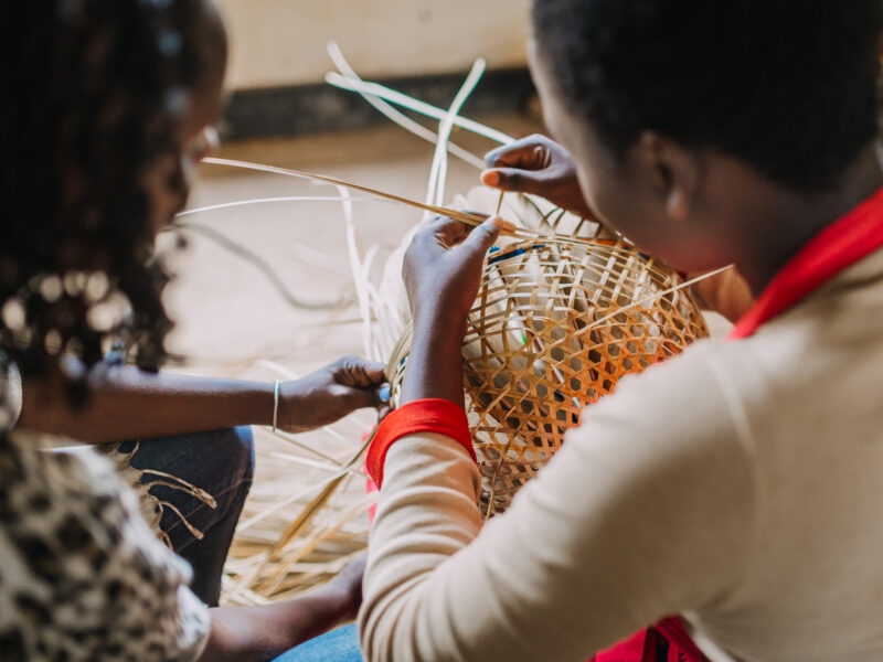 Over-the-shoulder view of two people hand-weaving a geometric pattern into a natural fiber basket.