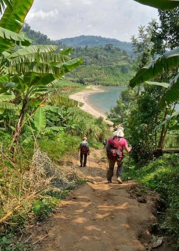 Two people hiking a dirt path surrounded by lush banana trees leading to a lakeside beach.