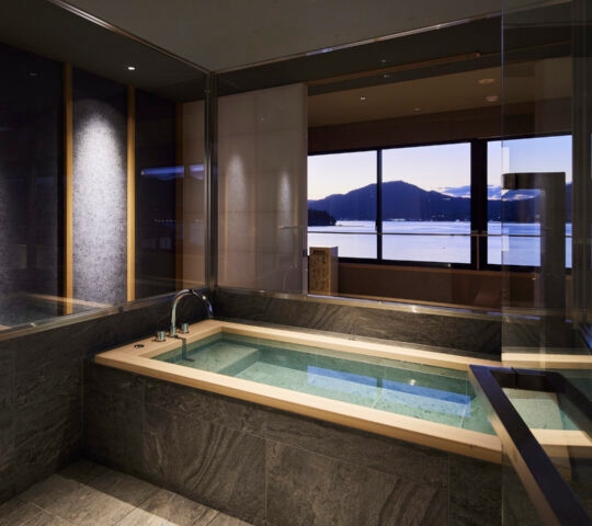 Interior of a corner bath with dual-aspect windows overlooking the sea and mountains with a purple sunset.