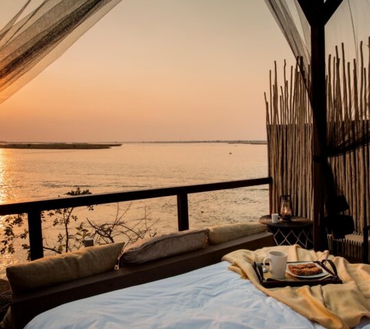 View from an outdoor bed with mosquito netting and a breakfast tray, looking over a wide river at sunrise.