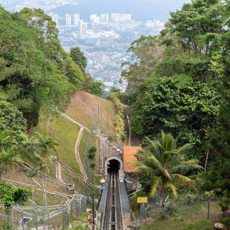 A railway line leading into a tunnel going through a hill surrounded by thick greenery with a city in the background in Asia