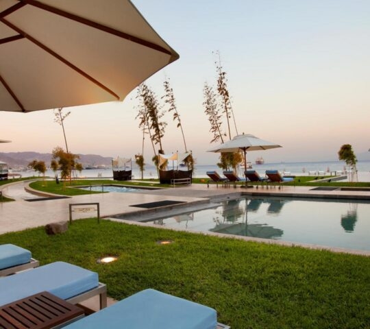 Blue lounge chairs and an umbrella by an outdoor pool at Kempinski Hotel Aqaba, overlooking the sea.
