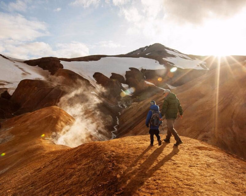 A man and a child hike along a volcanic mountain ridge in Iceland with steam rising from vents in the rocky ground.
