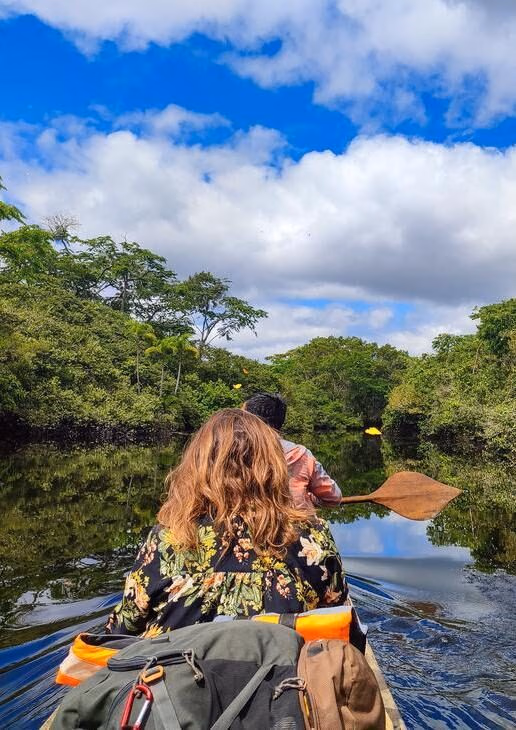 A person in a floral shirt paddles a canoe through a river in the Peruvian Amazon surrounded by lush green rainforest.