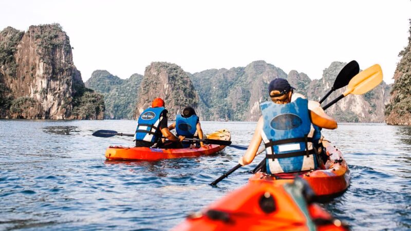 People wearing blue life jackets paddle orange kayaks through calm water surrounded by tall, rocky limestone cliffs.