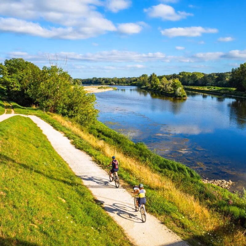 Two cyclists ride bikes on a trail beside a wide, calm river in the Loire Valley, France, surrounded by greenery.