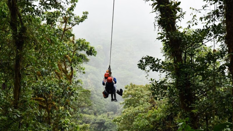 A group wearing red helmets zips along a cable suspended through the dense, misty canopy of a Costa Rican forest.
