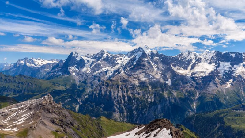 Panoramic view of a rugged, snow-capped Swiss mountain range rising above deep green valleys under a cloudy blue sky.