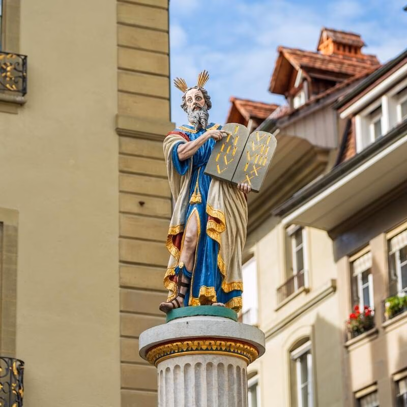 A vibrant painted statue of a bearded man holding stone tablets with Roman numerals on a public fountain column.