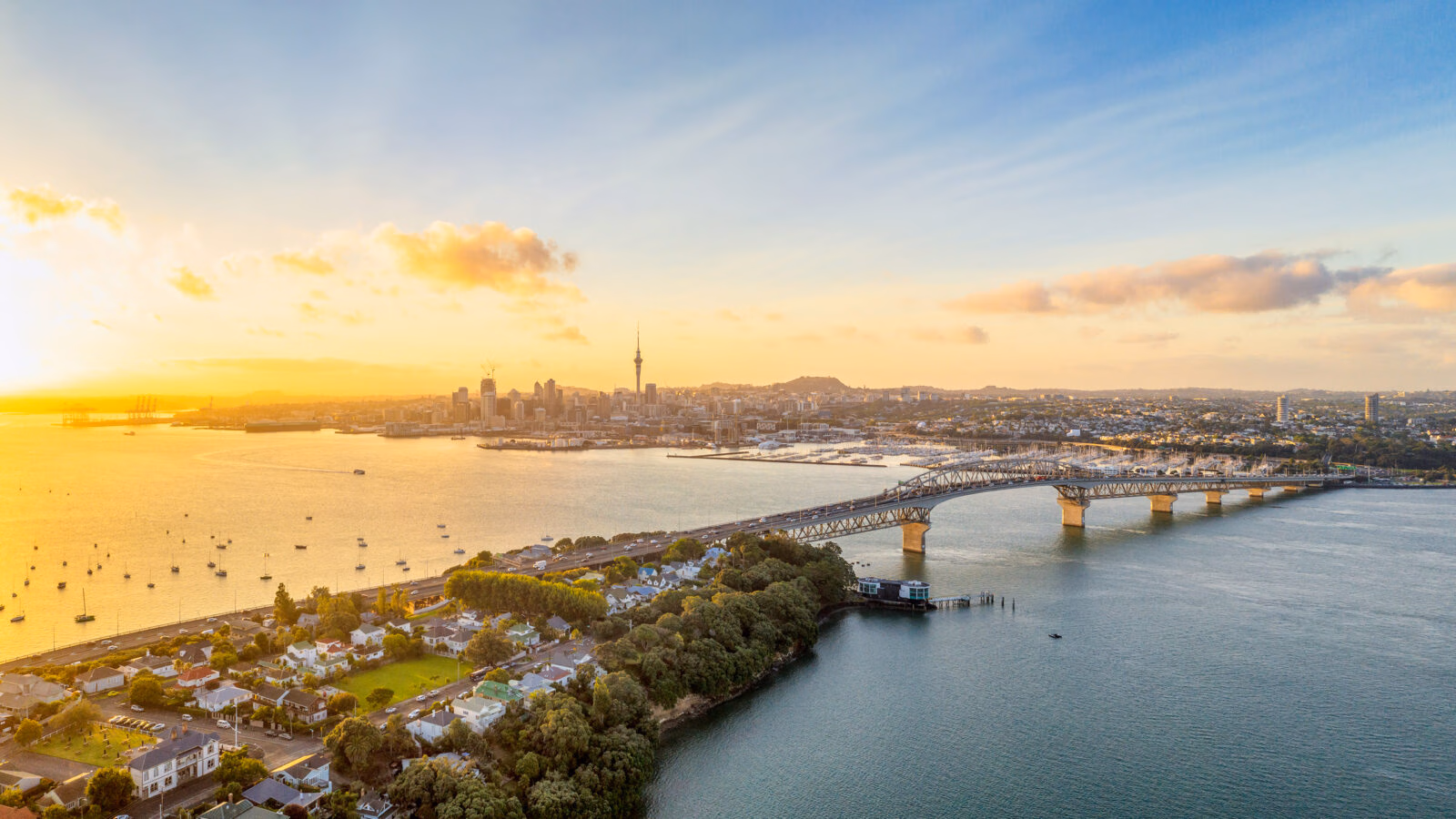 Aerial view of the Auckland skyline and Harbour Bridge at sunset during a family-trip, with boats on the golden water.