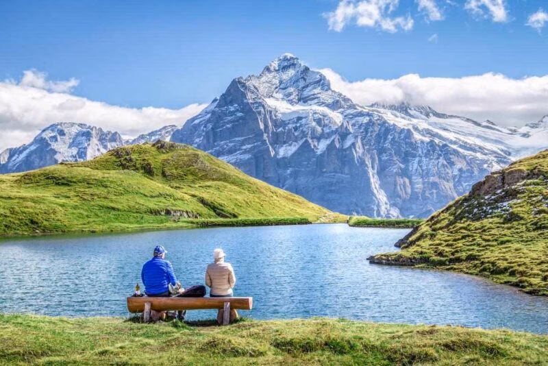 Two people sitting on a rustic bench looking toward a mountain lake and a massive snow-capped peak in Switzerland.