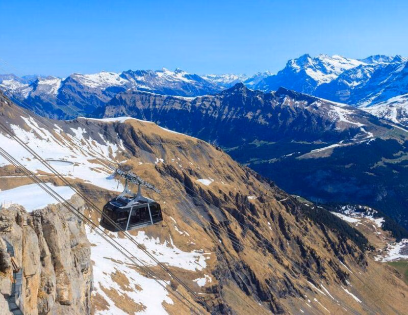 A modern cable car hanging from lines over a steep rocky cliff with vast mountain ranges stretching into the distance.